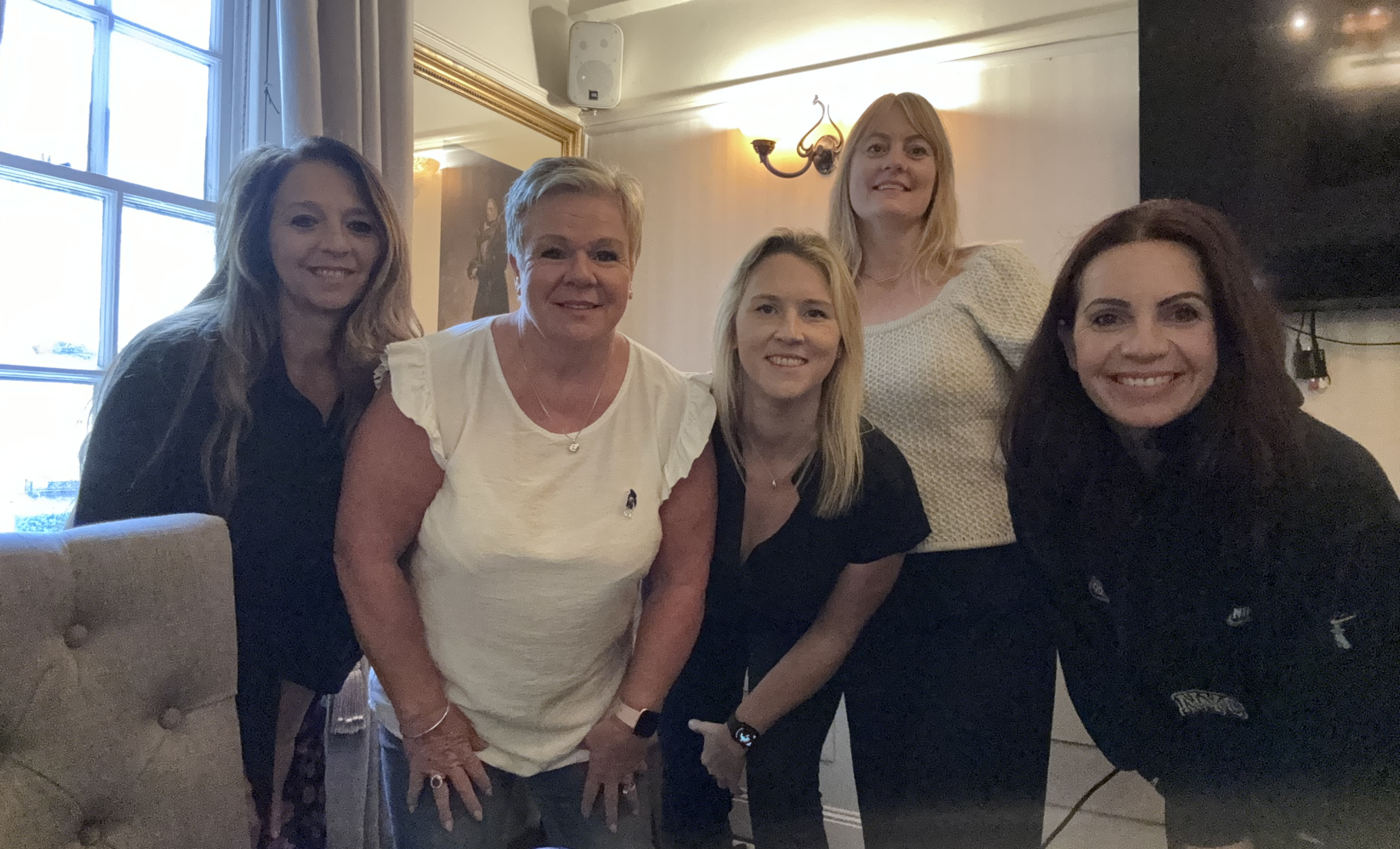 A group of 5 ladies stood posing for a picture. Three of the women have blonde hair and two women are brunette. All women are visibly smiling.